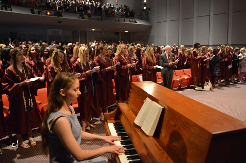 A woman plays piano with graduates in the background.