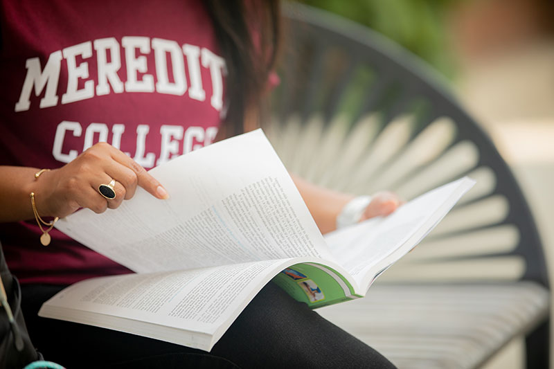Student in burgundy Meredith College sweatshirt sitting on a bench outside flipping through textbook.