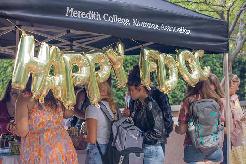 A large tailgate tent that says "Meredith College Alumnae Association" and balloons that say "Happy FDOC" with students getting goodies.