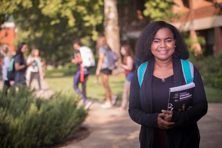 Marie Johnson Business major standing with book in hand and students walking in the distance.