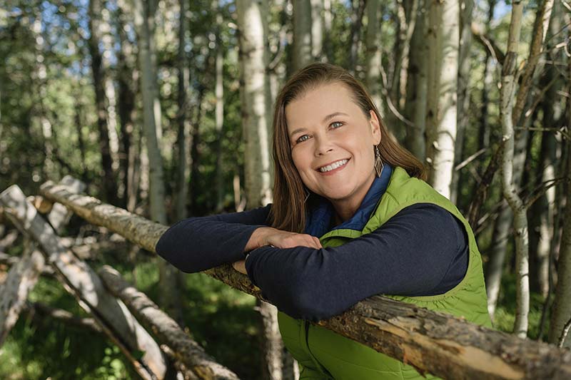 Heather White leaning against fallen tree branch in wooded area.